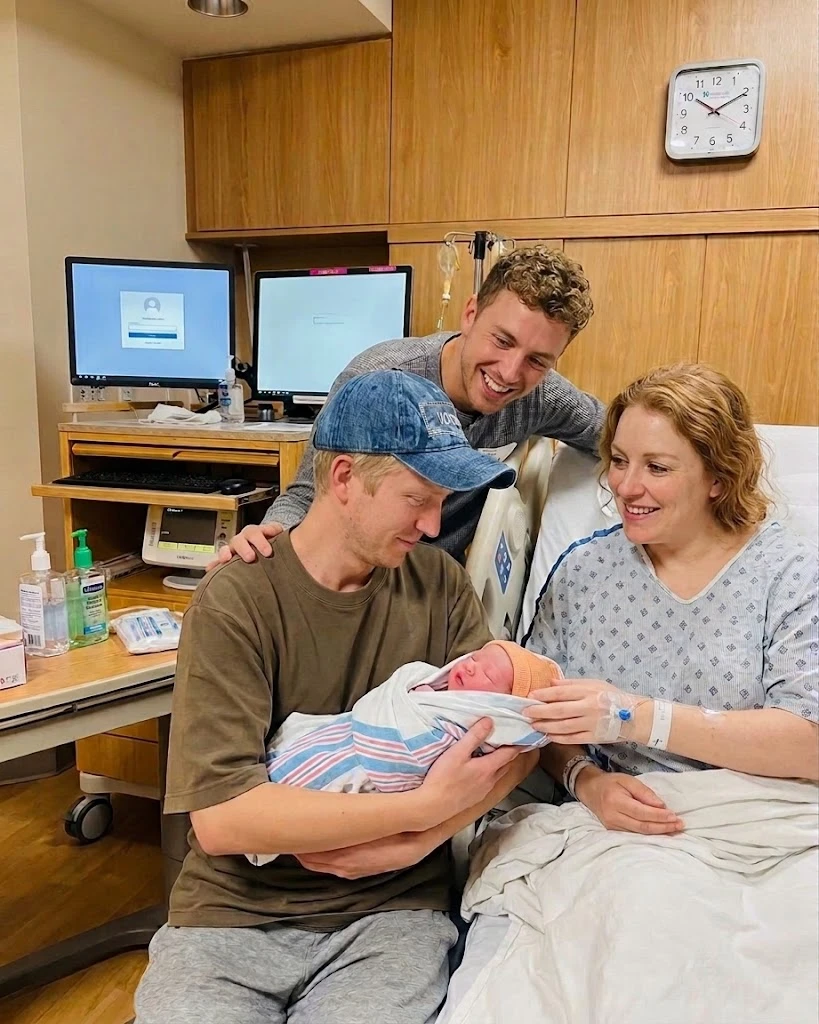 A man cradles a swaddled newborn beside a woman in a hospital bed, with another man smiling behind them